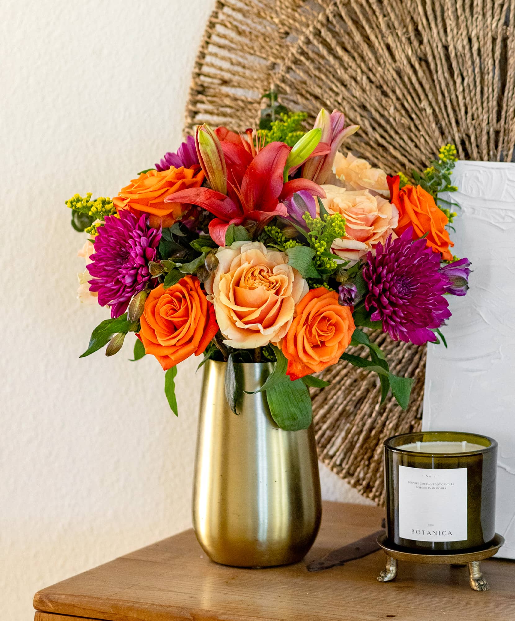 Vibrant floral arrangement with orange roses, lilies, and purple blooms in a sleek vase.